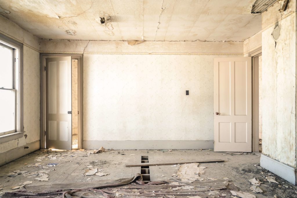 Deteriorated room with peeling wallpaper, damaged ceiling, and debris-covered floor.