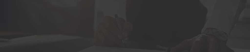 Person's hand signing document at desk, darkened overlay.