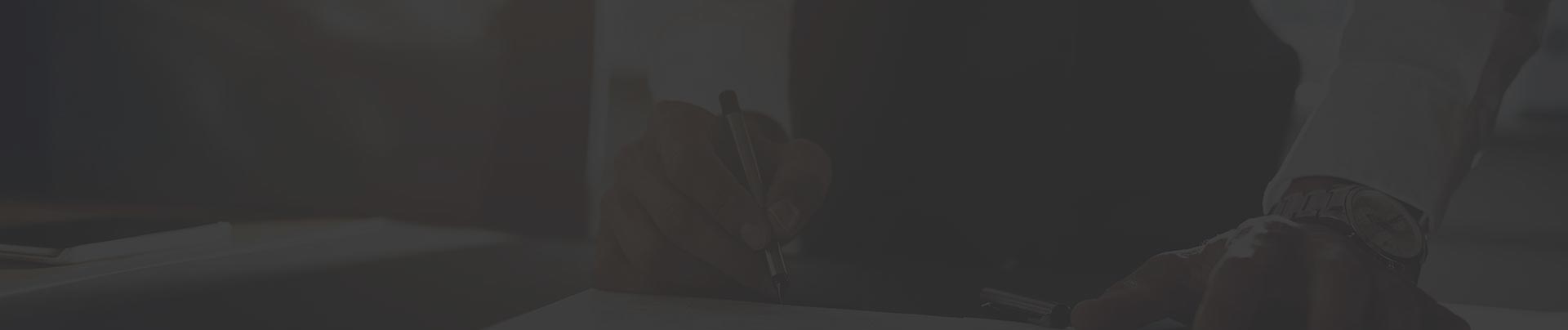 Person's hand signing document at desk, darkened overlay.