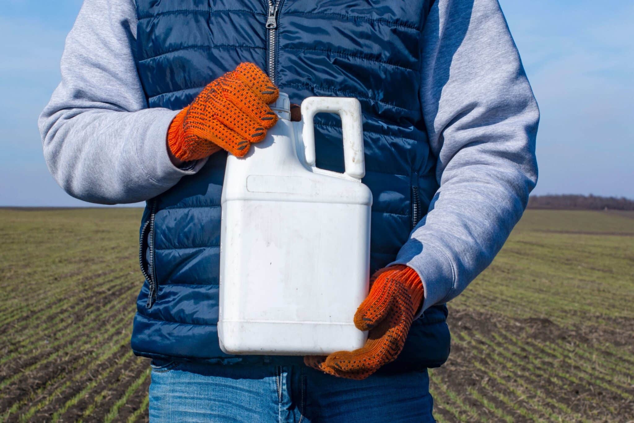 Farmer wearing gloves holding white jug of agricultural chemical in field.
