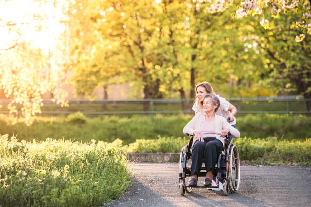 Woman pushing elderly woman in wheelchair along outdoor path at sunset.