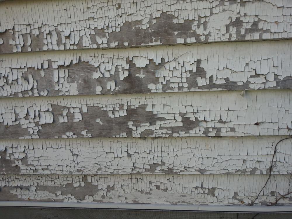 Peeling white paint on wood siding, exposing weathered wood underneath.