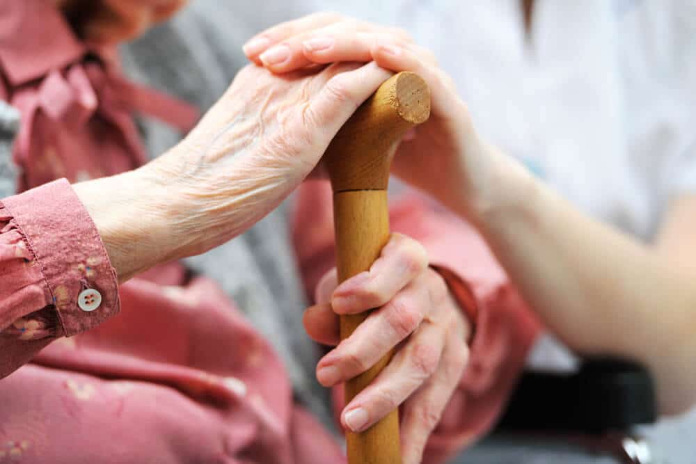Elderly woman's hands resting on wooden cane, caregiver's hand placed on top.