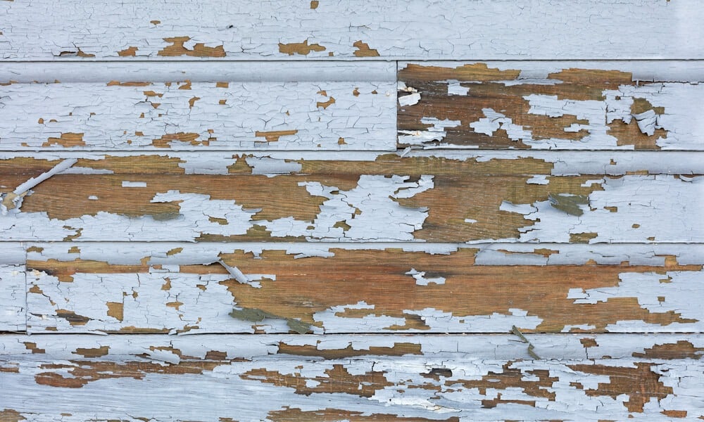 Peeling white paint on wood siding, revealing weathered wood grain underneath.