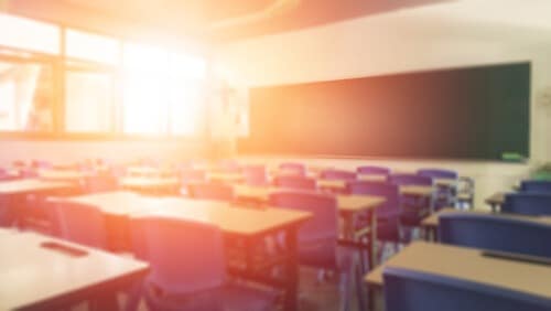 Empty classroom with rows of desks and chairs, sunlight streaming through windows.