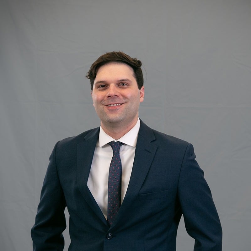 Fotografía profesional de Justin Lerche, Esq., sonriendo, vistiendo un traje azul marino con corbata estampada, sobre fondo gris.