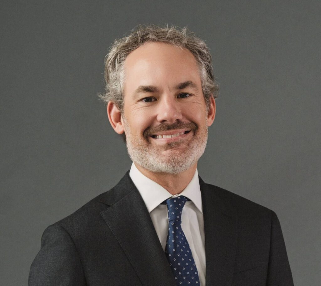 Fotografía profesional de Bryant McCulley, Socio Director, sonriendo, vistiendo un traje oscuro con corbata de lunares azul marino, sobre fondo gris.