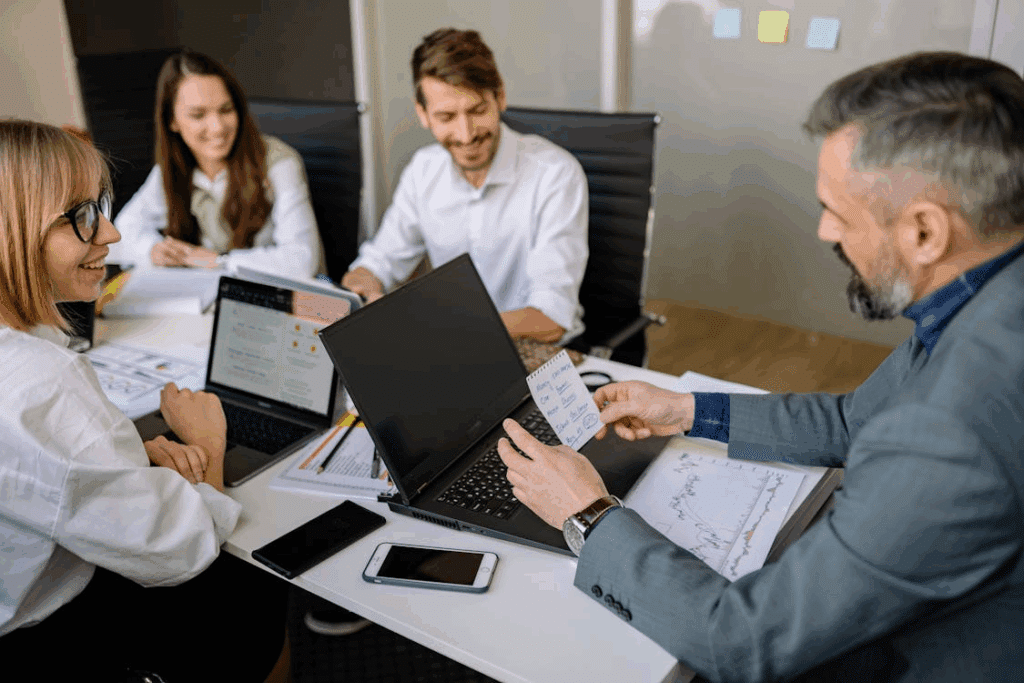 Four people around table with laptops, papers, and charts, discussing during business meeting.