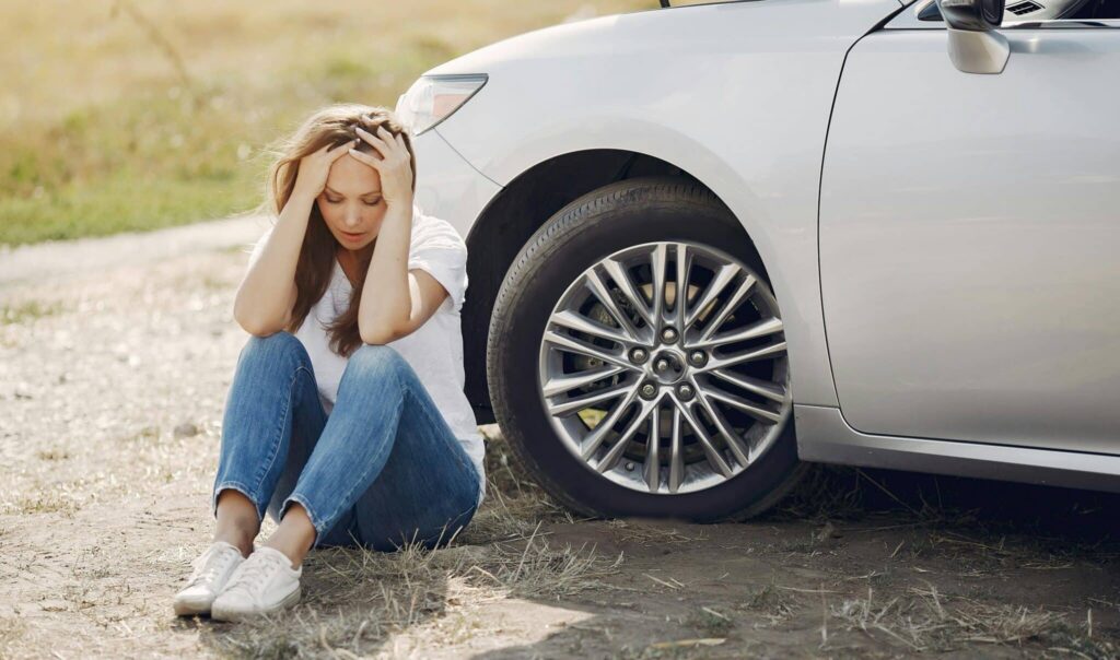 Woman sitting on ground beside car, head in hands, roadside breakdown
