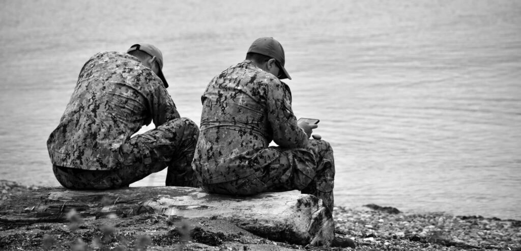 Two servicemembers in camouflage sitting on driftwood by water, black and white
