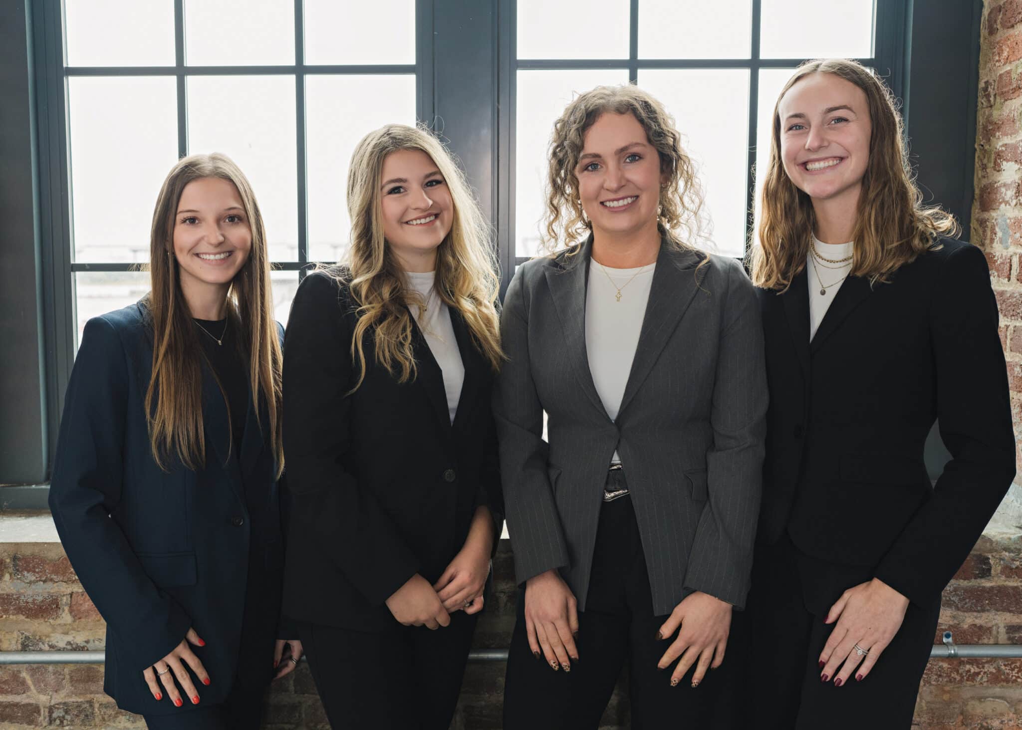 Four female staff members in business attire, group photo in front of brick wall and windows