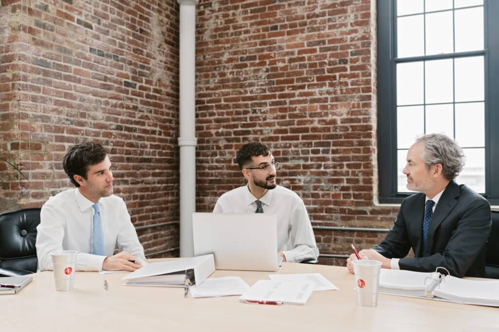 Three attorneys in meeting at conference table, brick wall office, reviewing documents and laptop
