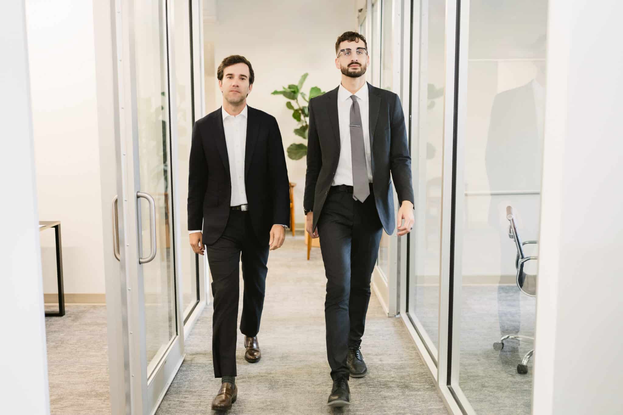 Two attorneys in suits walking through office hallway with glass-walled conference rooms