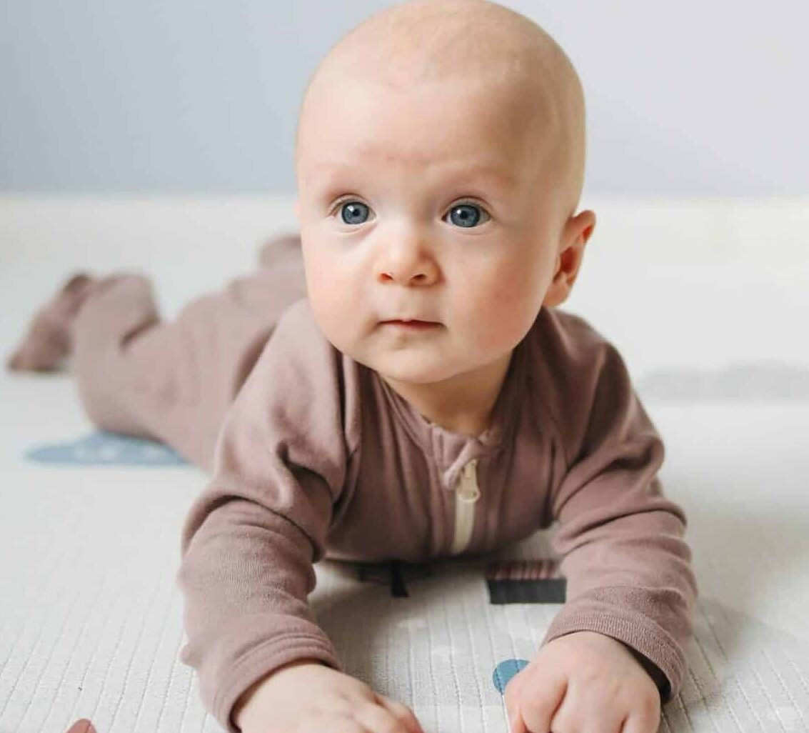 Infant with blue eyes lying on stomach on a textured play mat, propped up on forearms and looking toward the camera