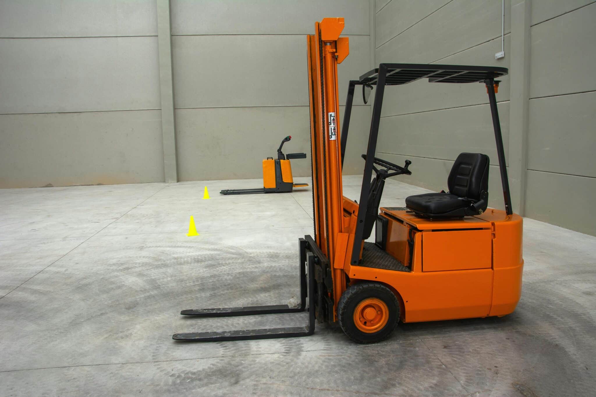 Orange forklift parked in warehouse, pallet jack and safety cones in background