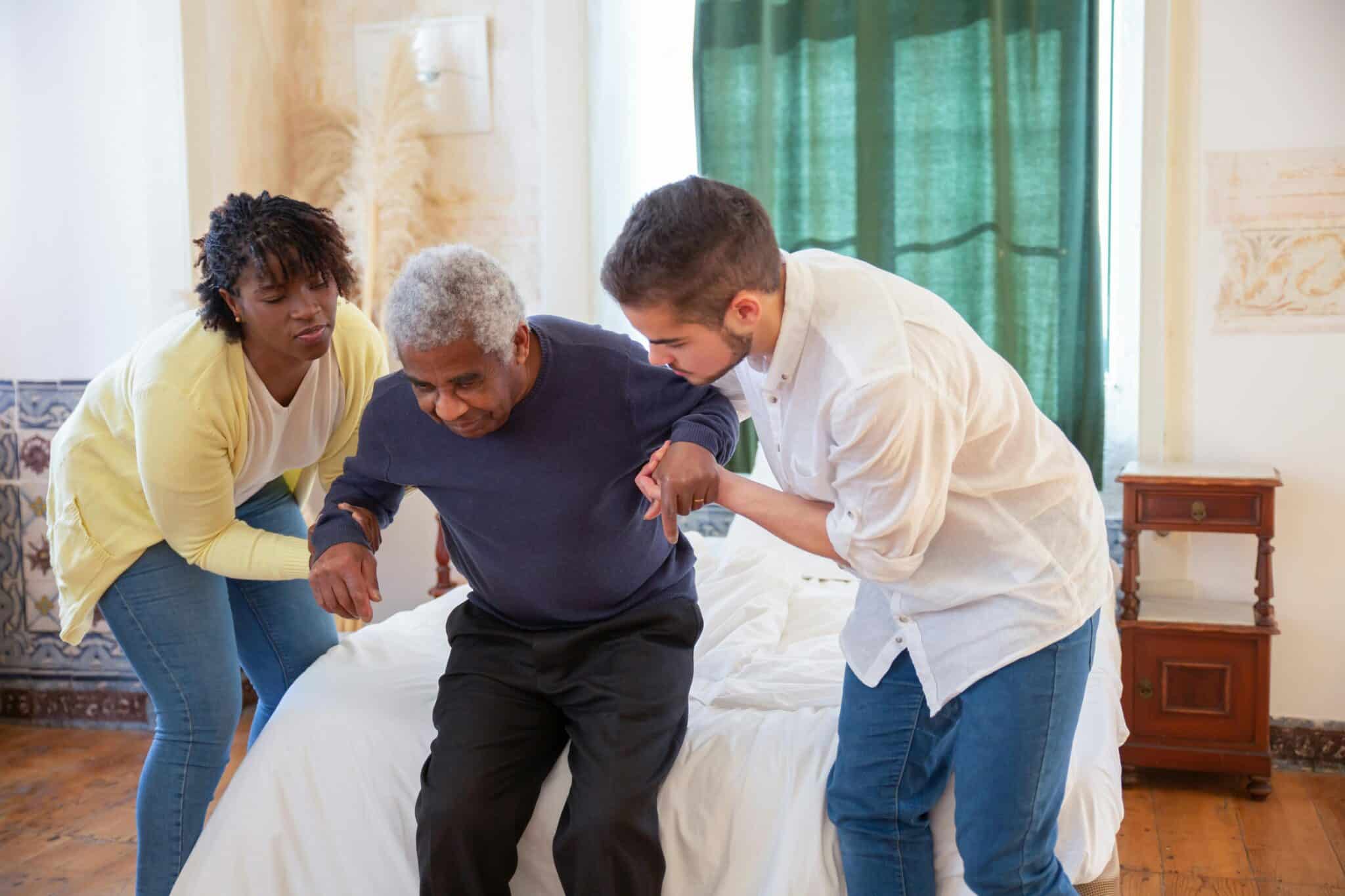 Two adult family members helping older man rise from bed