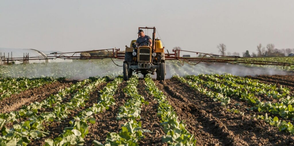 Agricultor conduciendo un tractor con aspersor de brazo, aplicando pesticida sobre hileras de cultivos Farmer driving tractor with boom sprayer applying pesticide over crop rows